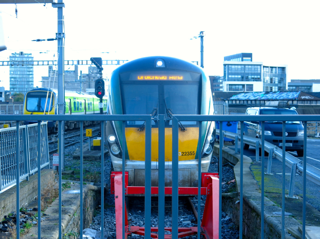 Zwei Züge auf dem Gleis mit einem Zaun im Vordergrund, Pfosten am Land, einer mit einer Signallampe, ein Auto auf der Straße rechts, Gebäude im Hintergrund und Himmel oben.
