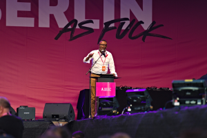 Ein Mann in einem weißen Hemd und braunen Hosen steht auf einer Bühne und hält ein Mikrofon, während er eine Menge anspricht. Im Hintergrund ist ein pinkfarbenes Banner mit der Aufschrift "Berlin as Fuck" zu sehen.