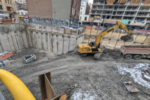 Eine Baustelle mit einem Bagger, Fahrzeugen, Holzgegenständen, einem gelben Rohr links, Gebäuden, Masten, Verkehrszeichen, Schildern, Bäumen und dem Himmel im Hintergrund.