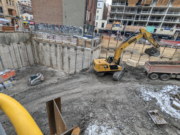 Eine Baustelle mit einem Bagger, Fahrzeugen, Holzgegenständen, einem gelben Rohr links, Gebäuden, Masten, Verkehrszeichen, Schildern, Bäumen und dem Himmel im Hintergrund.