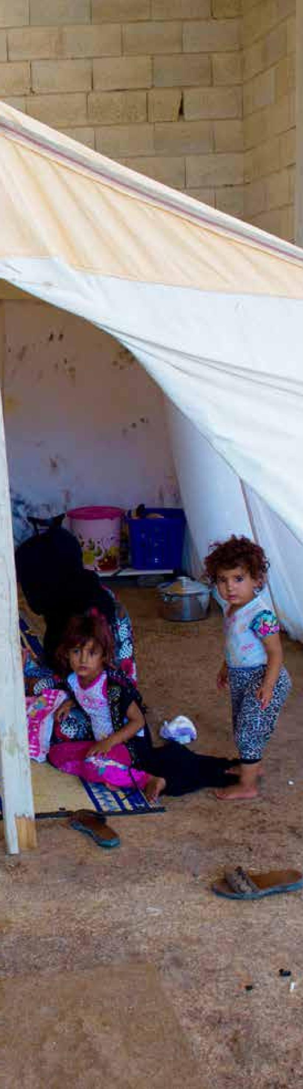 Drei Kinder in bunten Kleidern sitzen in einem Zelt auf einem Raumboden, mit verstreuten Gegenständen drumherum und einer Wand im Hintergrund.
