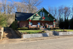 Ein gr├╝nes Haus mit einem Kranz vorne drauf, umgeben von Pflanzen, Gras, Treppen, einer Wand, einer Gruppe von B├Ąumen, einigen Dr├Ąhten und einem bew├Âlktem Himmel, das sich im Herzen der Stadt befindet und zur Miete verf├╝gbar ist.