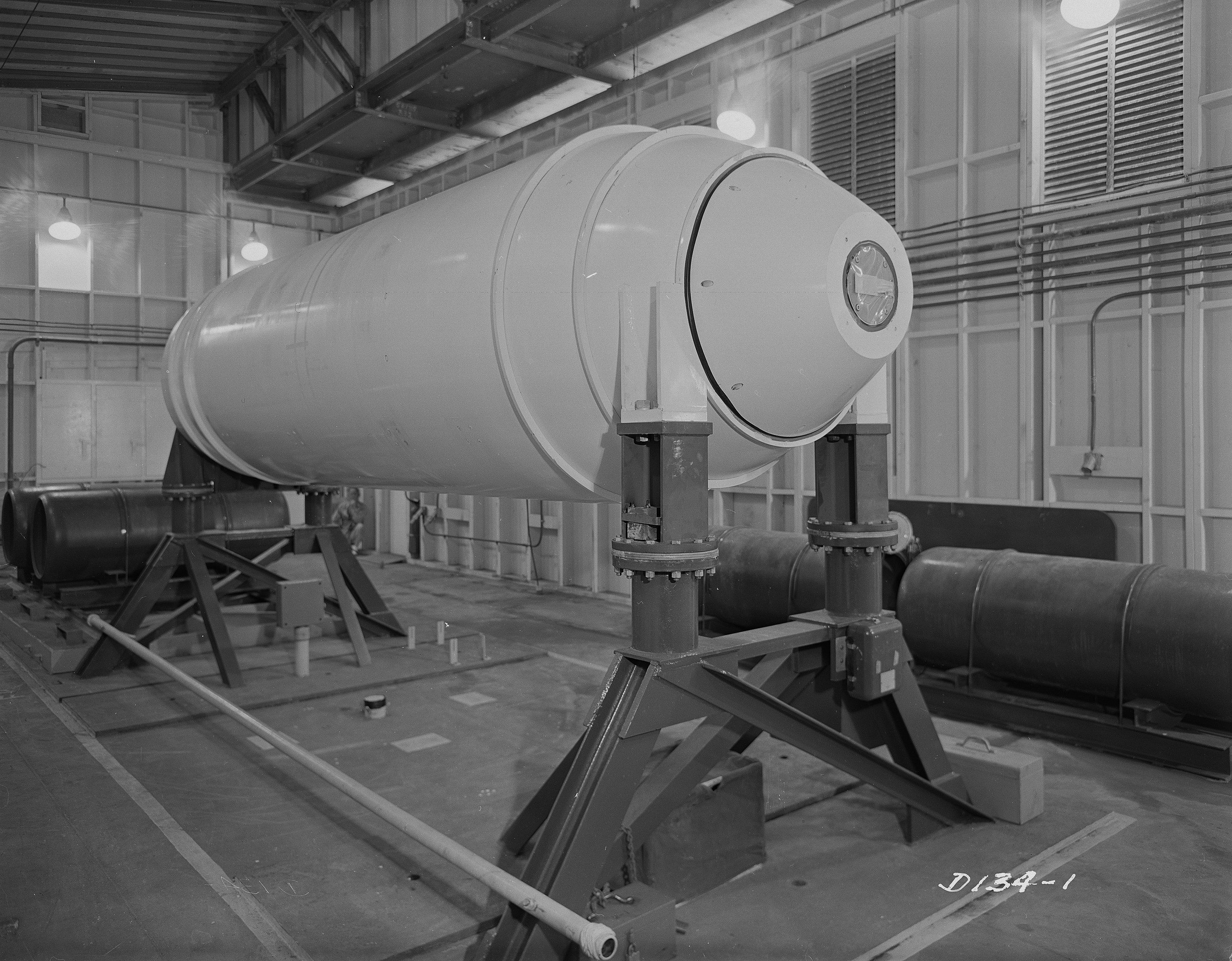Ein Schwarz-Weiß-Foto einer großen zylindrischen Wasserstoffbombe auf einem Ständer in einer Fabrik, mit Rohren, Lampen und einer Wand im Hintergrund.