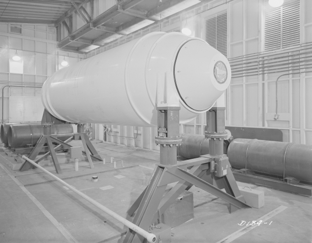 Ein Schwarz-Weiß-Foto einer großen zylindrischen Wasserstoffbombe auf einem Ständer in einer Fabrik, mit Rohren, Lampen und einer Wand im Hintergrund.