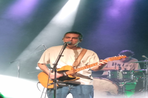 Ein Mann spielt eine E-Gitarre auf der Bühne, beleuchtet von Scheinwerfern von oben, während er in ein Mikrofon singt; ein anderer Mann spielt im Hintergrund Schlagzeug.