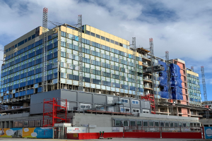 Großes Gebäude im Bau in einer städtischen Umgebung mit Glasfenstern, Metallrahmen, einer Treppe, einem Zaun, einem Schild, Pfählen, einer Straßenlaterne, Bäumen und einem bewölkten Himmel.