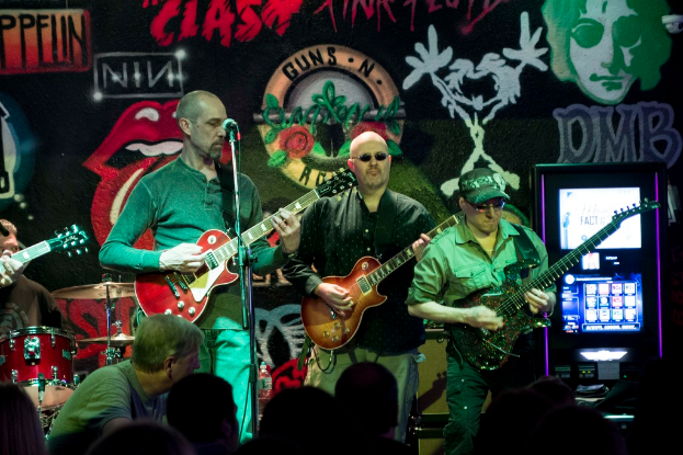 Drei Männer auf einer Bühne, die Gitarren spielen, mit einer Menge unten und einer gemalten Wand im Hintergrund.