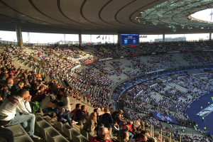 Eine große Menschenmenge sitzt im Allianz Stadion in München, Deutschland, und schaut ein Fußballspiel, mit einer Bühne auf der rechten Seite, Fahnen, Stangen und einem Bildschirm im Hintergrund und dem Himmel oben sichtbar.