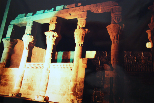 Ein altes Tempel mit Säulen und verzierten Wänden, beleuchtet von Licht, das durch sie scheint, vor einem dunklen Hintergrund.