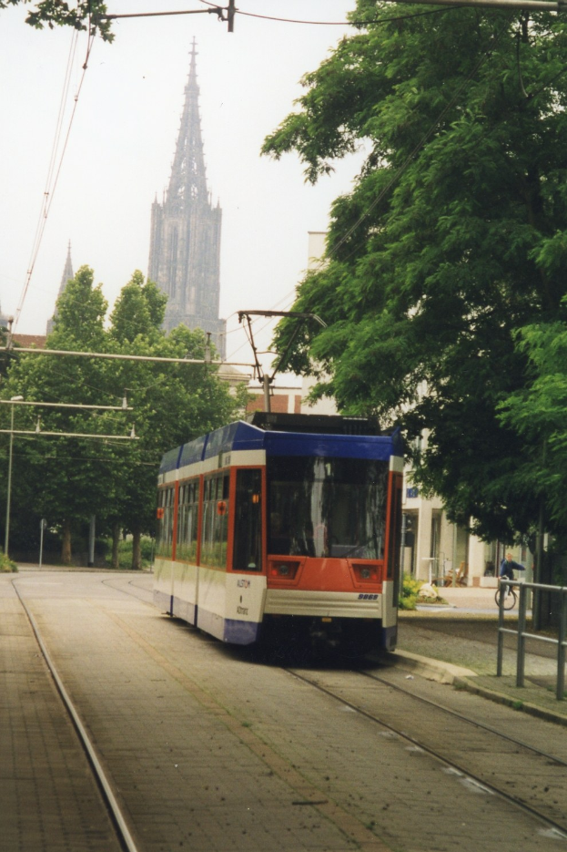 Rotes und weißes Zugfahrzeug auf Schienen neben einem hohen Gebäude, mit einer Person, die auf einem Gehweg neben den Schienen Fahrrad fährt, Bäume entlang der Schienen und Gebäude im Hintergrund unter einem klaren blauen Himmel.