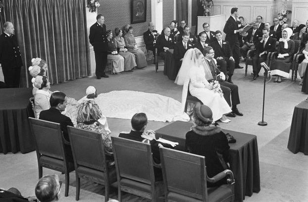 Schwarzes und weißes Foto einer Hochzeitszeremonie mit Menschen, die sitzen und stehen, einem Mann mit Mikrofon, einer Frau mit Blumenstrauß, einem Podium mit Mikrofon, Blumenschmuck, einem Vorhang, einem Rahmen an der Wand und einer Tür im Hintergrund.