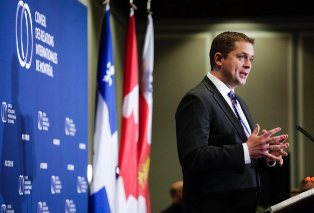 Der kanadische Premierminister steht an einem Rednerpult und hält eine Rede, umgeben von Flaggen und einem Banner, mit Menschen im Hintergrund.