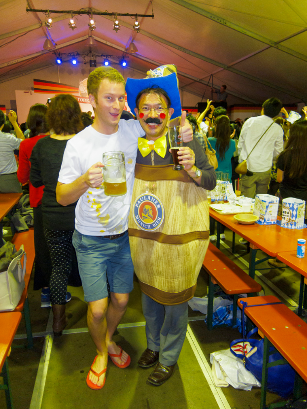 Zwei Personen stehen zusammen, eine hält ein Kind mit einem Bier, die andere trägt ein Dramakostüm, daneben ein Tisch mit Essen und eine Gruppe von Menschen dahinter.