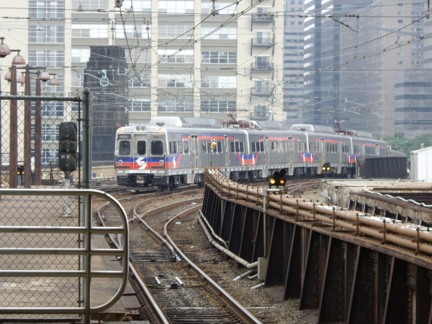 Ein Nahverkehrszug fährt auf Schienen neben hohen Gebäuden, mit verschiedenen Bahninfrastruktur- und Baumobjekten im Hintergrund.