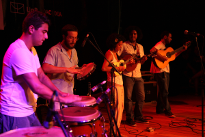 Eine Musikband auf der Bühne mit vier Mitgliedern: einem Schlagzeuger links, einem Gitarristen rechts, einem weiteren Gitarristen neben ihm und einer Person, die ein Instrument in der Mitte spielt, alle mit Mikrofonen und Kabeln auf dem Boden.