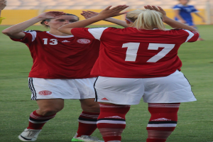 Zwei Frauen in roten T-Shirts und weißen Shorts klatschen auf einem Fußballfeld mit einer Person in einem blauen Kleid und einem gelben Brett im Hintergrund.