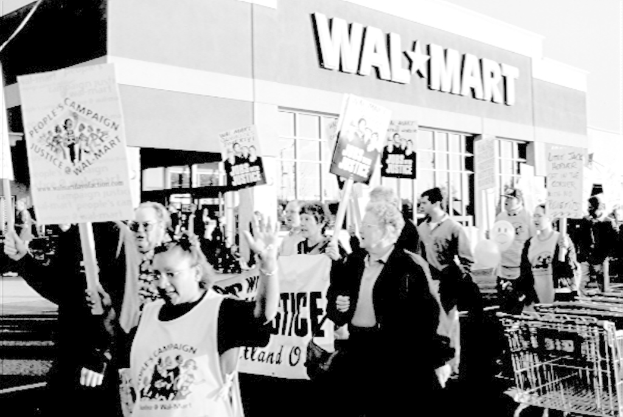 Ein Schwarz-Weiß-Bild von protestierenden Menschen mit Schildern und einem Einkaufszentrum im Hintergrund.