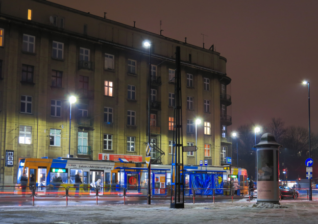 Stadtstraße bei Nacht mit einem angehaltenen Bus, Menschen an einer Bushaltestelle, Fahrzeuge, ein Metallzaun, Pfosten, Schilder, Straßenlaternen, ein Gebäude mit Fenstern, Bäume und eine bewölkte Nacht.