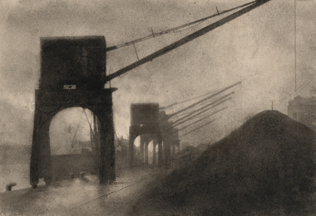 Ein Schwarz-Weiß-Foto einer Brücke im Bau, mit Gebäuden, Polen und Drähten im Hintergrund.