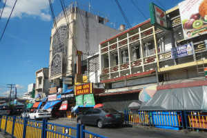 Eine belebte Stadtstraße mit Autos, hohen Gebäuden, Schildern, Regenschirmen, einem Metallzaun, Strommasten mit Drähten und einem bewölkten Himmel.