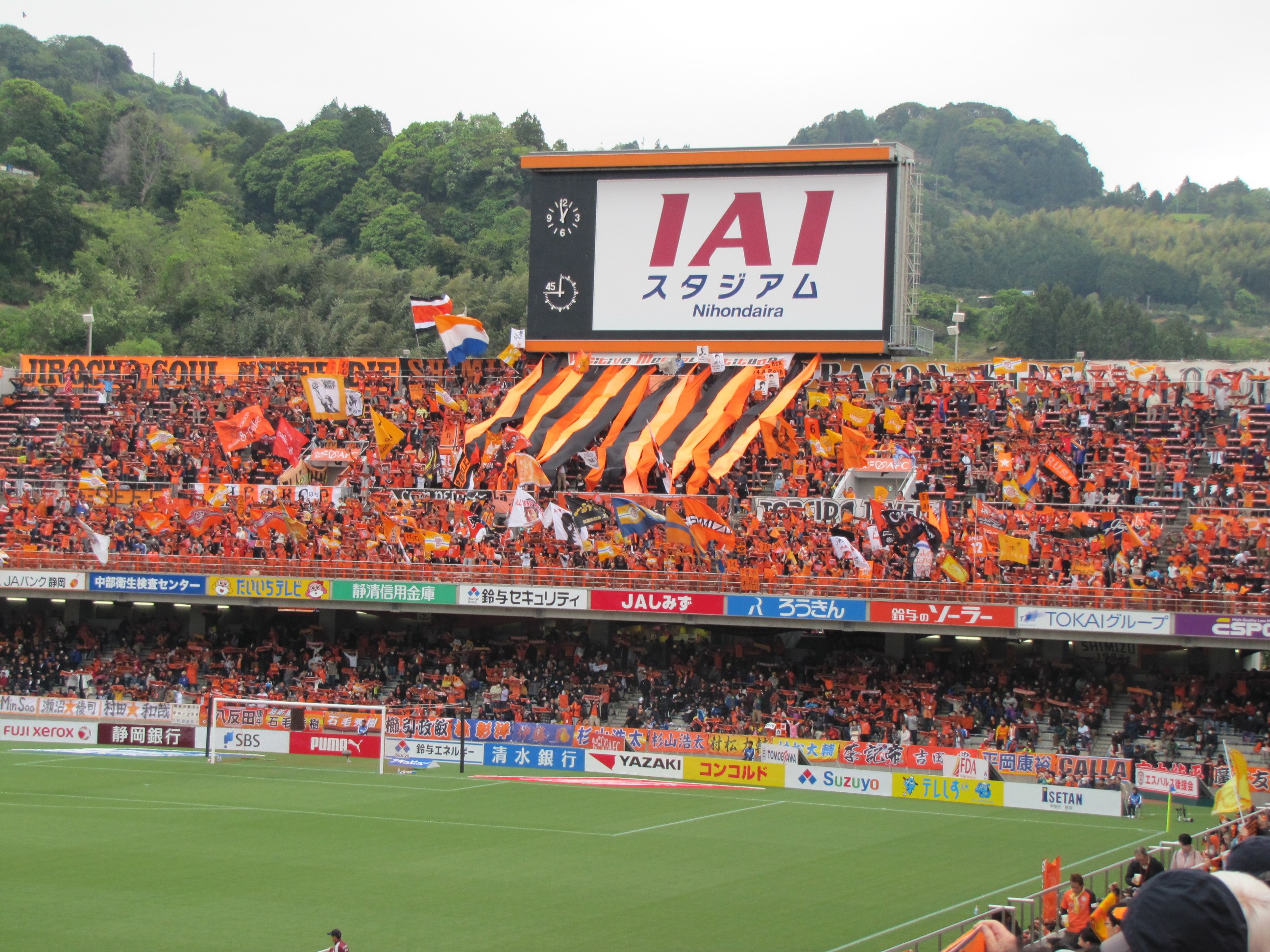 Ein Fußballspiel wird in einem Stadion mit einer großen Menge, grünem Gras, einem Torpfosten, Bannern, Fahnen, einem großen Bildschirm, Bäumen und einem klaren blauen Himmel gespielt.