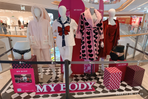 Eine Schaufensterauslage mit Schaufensterpuppen auf einem schwarz-weißen Schachbrettboden, umgeben von Kartons, Kleidung, Beschilderung und einer Gruppe von Menschen, mit einer Tafel, die "My Body" anzeigt.