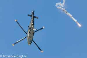 Ein militärisches Hubschrauber in der Luft, das eine Rakete hält, die Rauch und Feuer ausstößt, mit Text in der linken unteren Ecke.