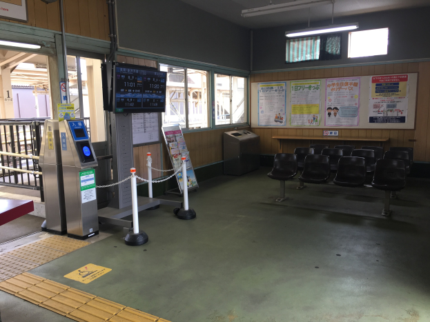 Ein belebter Bahnhofseingang mit Reihen von Stühlen, einem Service-Schalter, Ticketautomaten, Kettenzaunpfählen, Informationsanzeigen, Wandplakaten, Deckenbeleuchtung und Geländern.