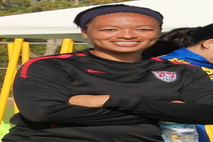 Zwei Frauen stehen auf einem Feld, eine hält eine Wasserflasche, mit einem Zelt, gelben Pfosten, Bäumen und einem klaren blauen Himmel im Hintergrund.