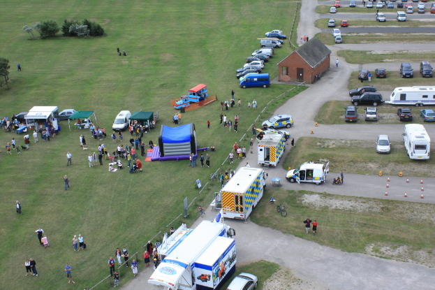 Eine Vogelperspektive eines Parkplatzes mit vielen Fahrzeugen, einigen Menschen in der Nähe von Zelten, einer zentralen Hütte und einigen Bäumen auf der linken Seite.