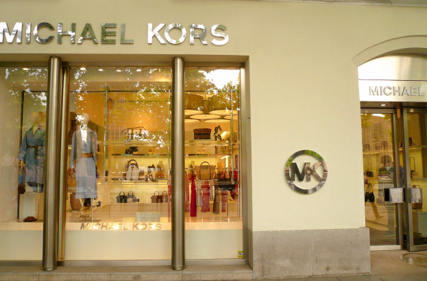 Ein Michael Kors Store in New York City mit Glaswänden und -türen, der Schaufensterpuppen in Kleidern und Regalen mit Handtaschen zeigt, mit einem Schild, das eine bevorstehende Eröffnung anzeigt.