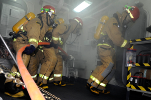 Feuerwehrleute in Schutzausrütstung, darunter Helme und Sauerstoffflaschen, arbeiten an einem Feuerwehrauto, mit einem Gestell voller verschiedener Gegenstände auf der rechten Seite und Rohren, Lichtern und anderen Gegenständen im Hintergrund.