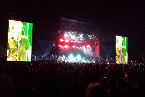 Eine Konzertszene mit einer Menschenmenge unten, einem großen Bildschirm links und einem kleineren Bildschirm rechts.