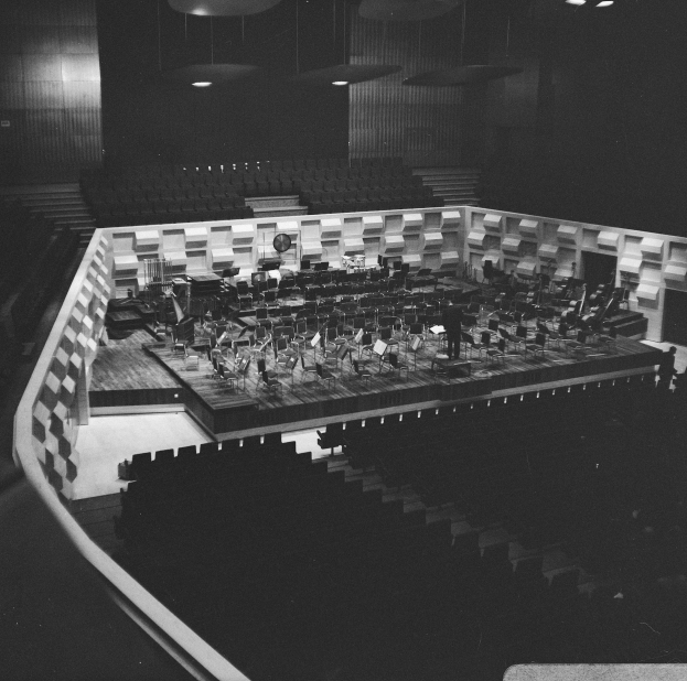 Ein Schwarz-Weiß-Foto eines großen Auditoriums mit einer Bühne in der Mitte, umgeben von Stühlen, auf der Musiker Instrumente spielen.