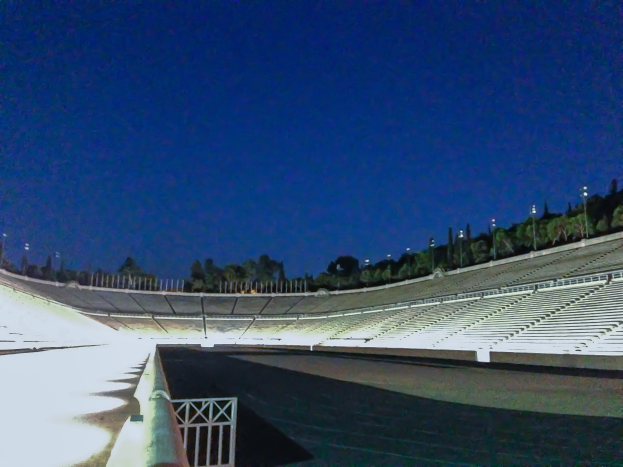 Olympiastadion bei Nacht, beleuchtet mit Stadionlichtern und sichtbaren Sternen am Himmel, umgeben von Bäumen und Pfählen, mit dem Boden unten sichtbar.