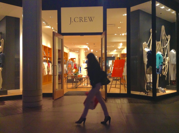 Ein Einkaufszentrum mit Glaswänden und -türen, das Puppen, Tische, hängende Kleider und Schränke zeigt, mit einer Frau, die daneben geht.