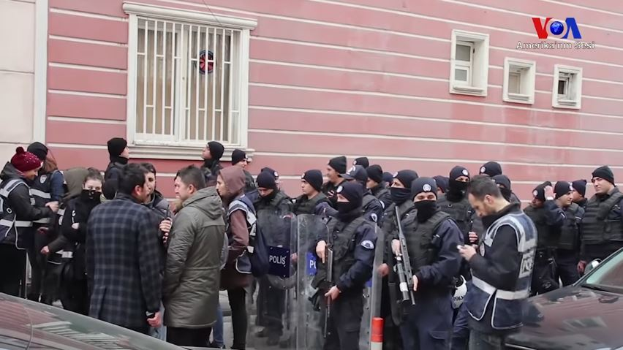 Eine Gruppe von Menschen, einige bewaffnet und mit Kappen bekleidet, steht vor einem Gebäude mit Fenstern, versammelt, um gegen eine Regierungsentscheidung zu protestieren, mit Fahrzeugen und einem Verkehrskegel im Vordergrund und einem Wasserzeichen auf dem Bild.