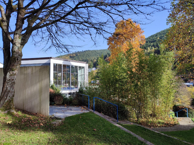 Das Gebäude der Universität Stuttgart mit einem Baum davor, umgeben von Gras, trockenen Blättern, Pflanzen und einem Geländer, mit Hügeln und einem klaren blauen Himmel im Hintergrund.