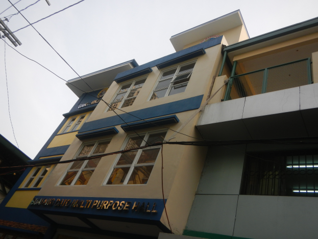 Ein Mehrzweckgeb├Ąude mit einer blauen und gelben Fassade, Glasfenstern, einem Namensschild, einem elektrischen Mast mit Dr├Ąhten und einem klaren blauen Himmel im Hintergrund.