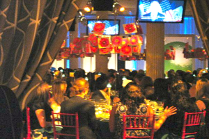 Eine Gruppe von Menschen an Tischen mit Essen, Fernsehern, Lampen, hängenden Papieren und Vorhängen in einer scheinbar festlichen Umgebung.