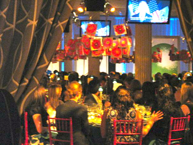 Eine Gruppe von Menschen an Tischen mit Essen, Fernsehern, Lampen, hängenden Papieren und Vorhängen in einer scheinbar festlichen Umgebung.