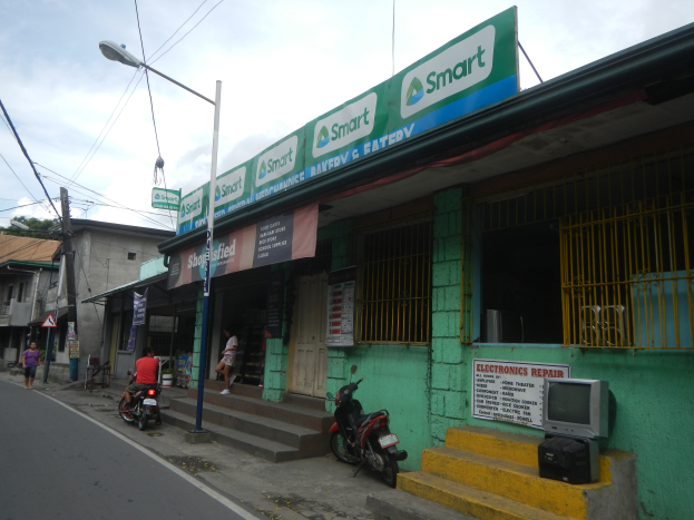 Ein grünes Gebäude mit einem Schild, umgeben von Motorrädern, Fußgängern, Straßenlaternen, Strommasten, Kabeln, Bäumen und einem bewölkten Himmel.