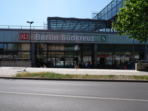 Ein gro├če Glaswand-Bahnhof namens "Berlin Südkreuz" in Berlin, Deutschland, mit Stra├čenlaternen, Lichtern, Fahrzeugen, Fü├čgängern, Fahrr├Ądern, B├Ąumen und einem klaren blauen Himmel.
