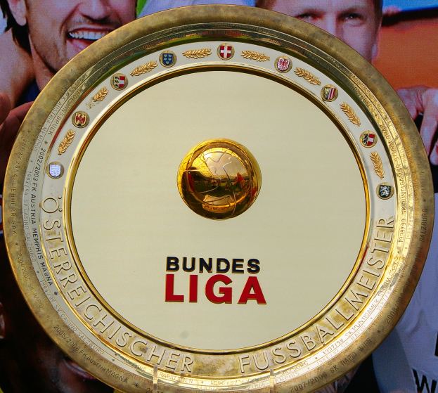 Ein Mann hält einen goldenen Schild mit der Aufschrift "Bundesliga" und im Hintergrund eine Fahne mit drei Personen.
