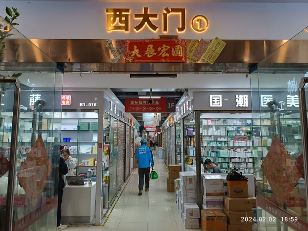 Innenansicht einer chinesischen Apotheke mit Glaswänden, Regalen voller Artikel, verstreuten Kartons auf dem Boden, einer Pflanze links und einer Namensschild oben.