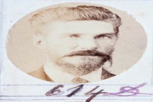 Ein altes Foto eines bärtigen Mannes mit Schnurrbart, der direkt in die Kamera schaut, mit leicht geöffneten Lippen, ordentlich gekämmtem Haar und einem gut gepflegten Bart.