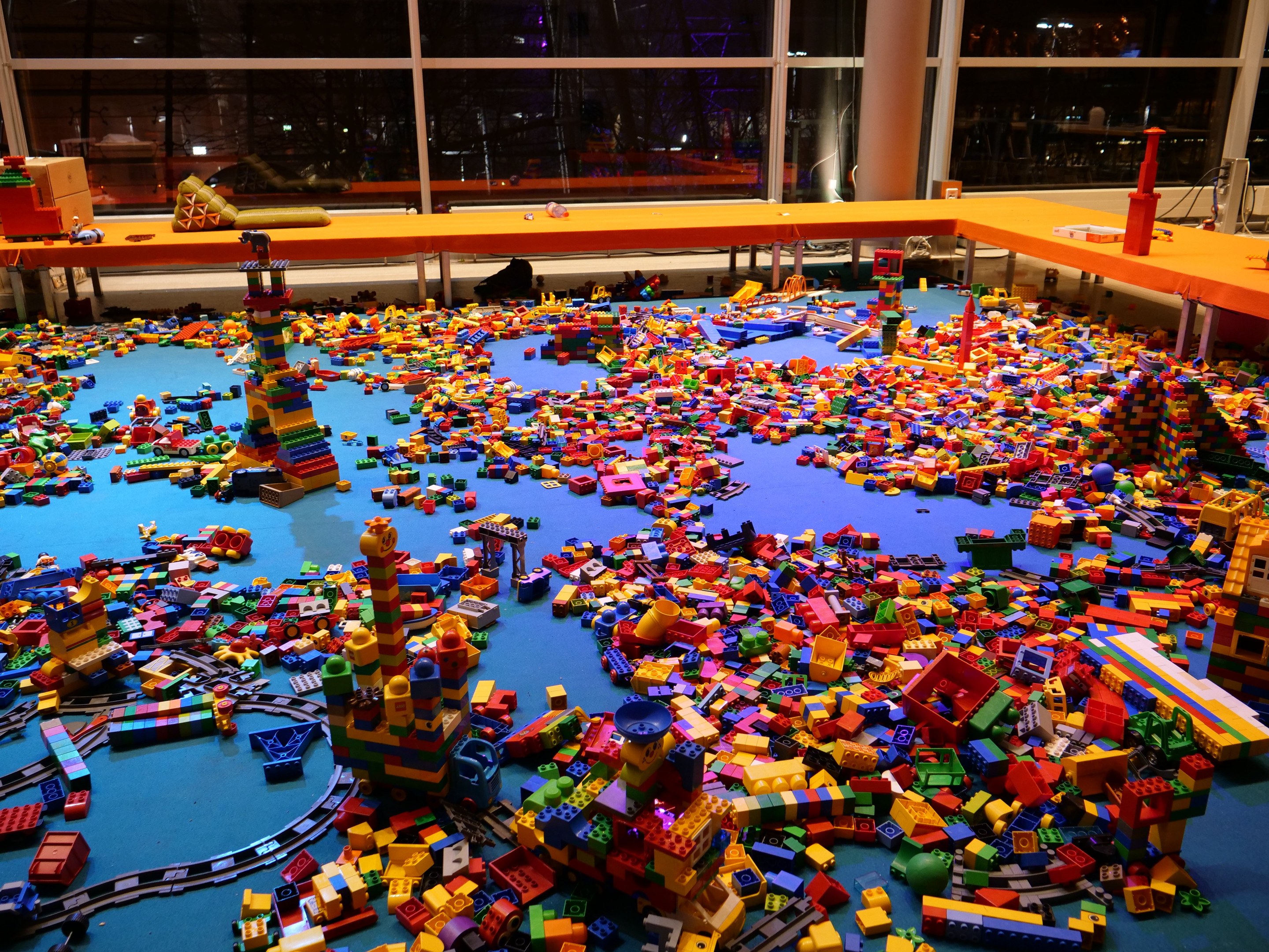 Ein geräumiger Raum mit farbigen Lego-Steinen auf dem Boden, umgeben von Tischen und Säulen, mit Blick durch Glasfenster auf Menschen und Ausstellungsstücke.