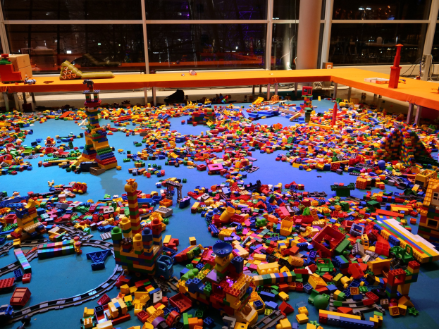Ein geräumiger Raum mit farbigen Lego-Steinen auf dem Boden, umgeben von Tischen und Säulen, mit Blick durch Glasfenster auf Menschen und Ausstellungsstücke.