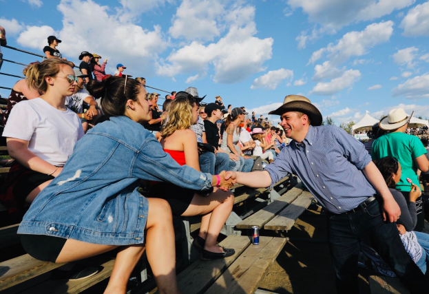 Ein Mann in einem blauen Hemd und schwarzen Hosen gibt einer Frau in unbekannter Kleidung die Hand, vor einem Publikum auf Bänken sitzend, mit weißen Zelten, Bäumen und einem blauen und weißen Himmel im Hintergrund.
