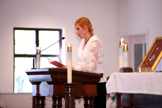 Eine Person in Kleidung steht an einem Podium mit einem Mikrofon, schaut auf ein Buch, mit einem tischgedeckten Tisch und einem Fenster im Hintergrund.